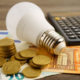 Eine Lampe liegt neben gestapelten Münzen und einem Taschenrechner – Symbolmotiv für Energiekosten und die Planung des Haushaltsbudgets.