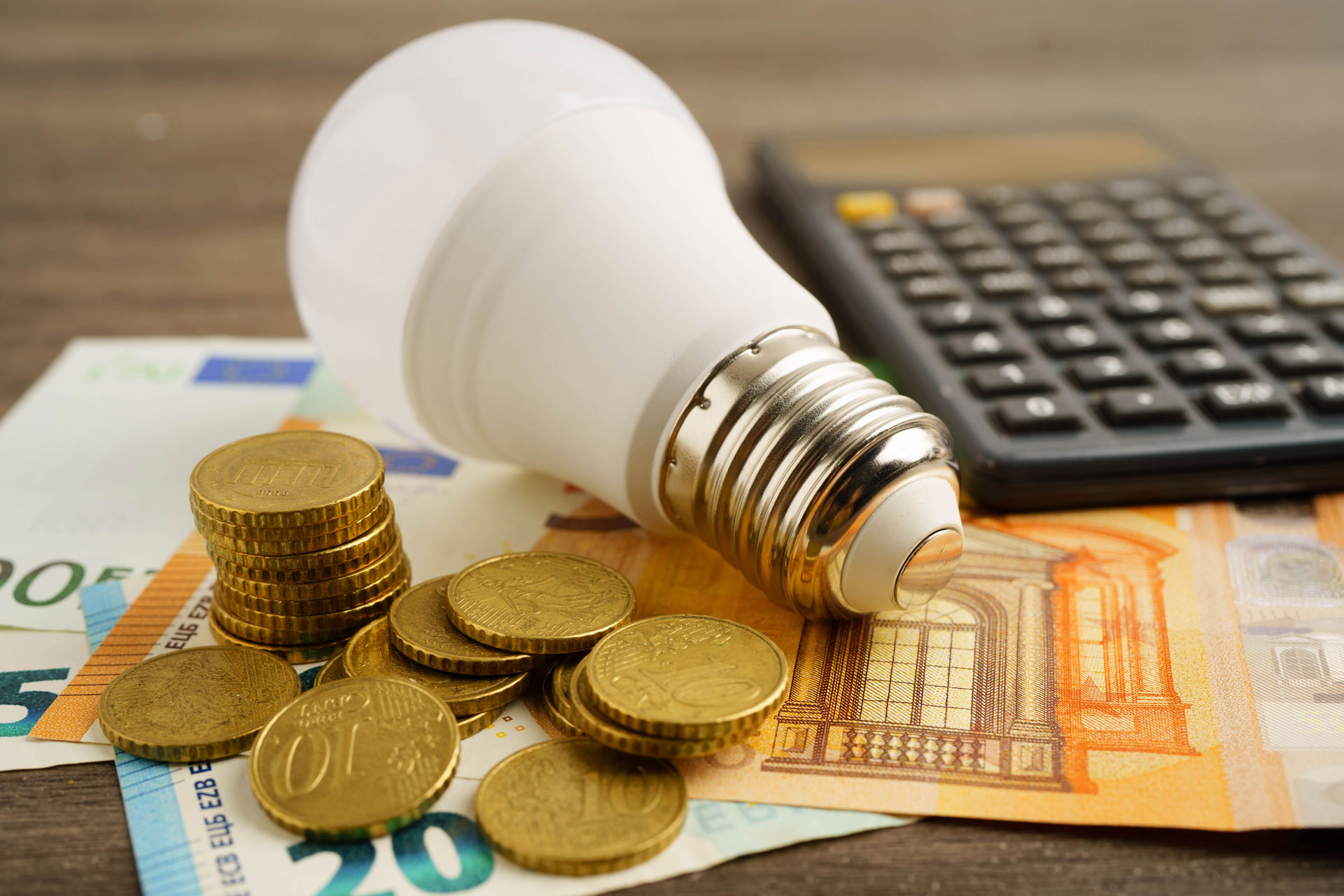 Eine Lampe liegt neben gestapelten Münzen und einem Taschenrechner – Symbolmotiv für Energiekosten und die Planung des Haushaltsbudgets.