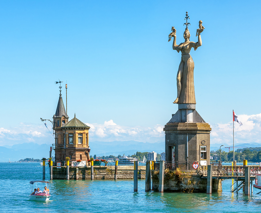 Die Imperia-Statue an der Hafeneinfahrt von Konstanz am Bodensee, mit dem Hafenturm und weiteren Gebäuden im Hintergrund.
